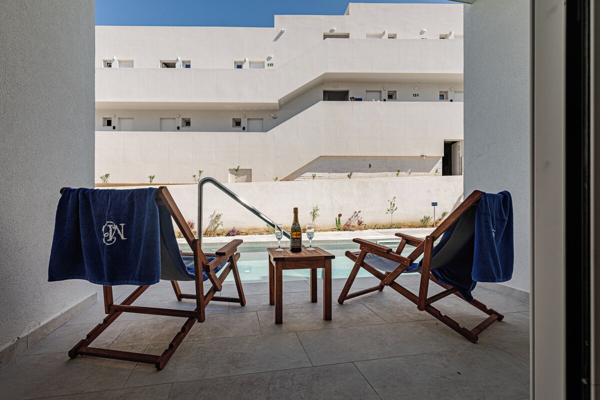 Un espace aménagé au bord de la piscine, avec des chaises longues et une petite table, une bouteille de vin, et une vue sur un bâtiment moderne.