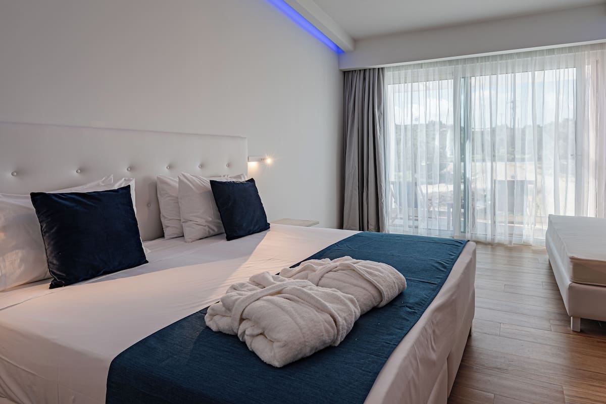 A modern hotel room with a large bed, a bedside table, and a view of the outdoors through sheer curtains.
