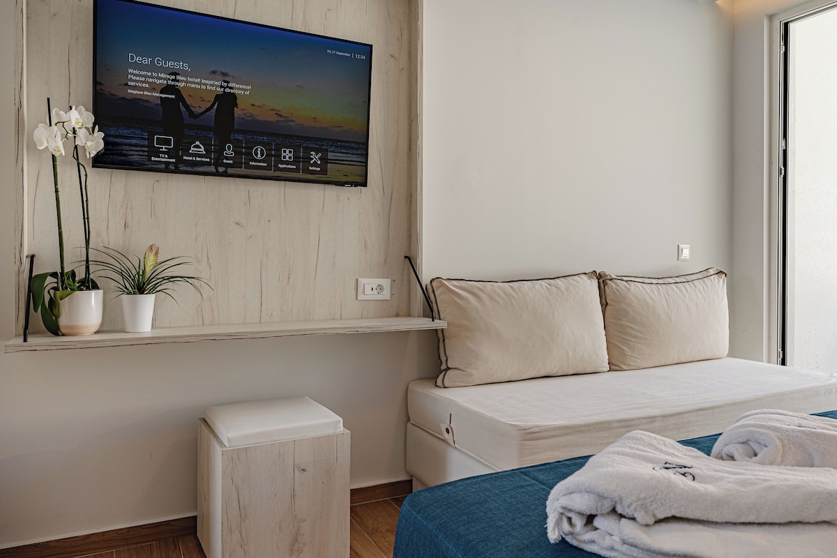 A hotel room with a bed, a TV mounted on the wall displaying a welcome message, a small table with a potted plant, and a window with a view of the sea.