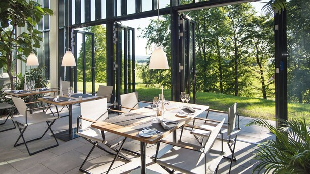 Un área de comedor al aire libre con mesas y sillas, ventanas amplias y follaje visible a través del vidrio.