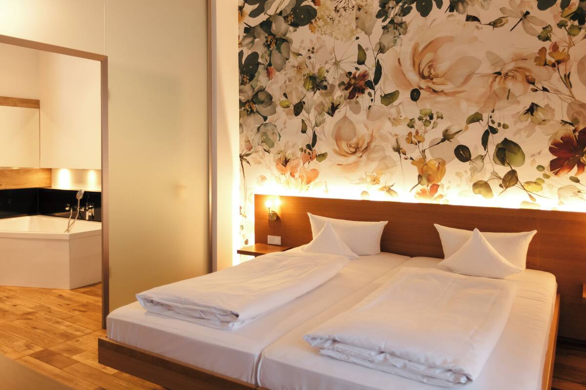 A hotel room with a large bed, a wooden headboard, a floral wallpaper, a mirror, and a bathroom with a sink and a wooden floor.