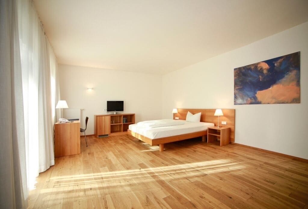 A modern hotel room with a wooden floor, a bed with white linens, a desk with a lamp, a television, and a painting on the wall.