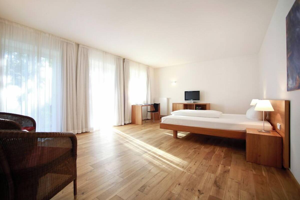 A modern hotel room with a wooden floor, a bed, a desk with a chair, a television, and a window with curtains.
