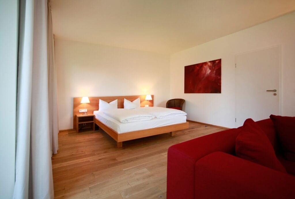 A hotel room with a wooden floor, a bed with white linens, a red sofa, and a red abstract painting on the wall.