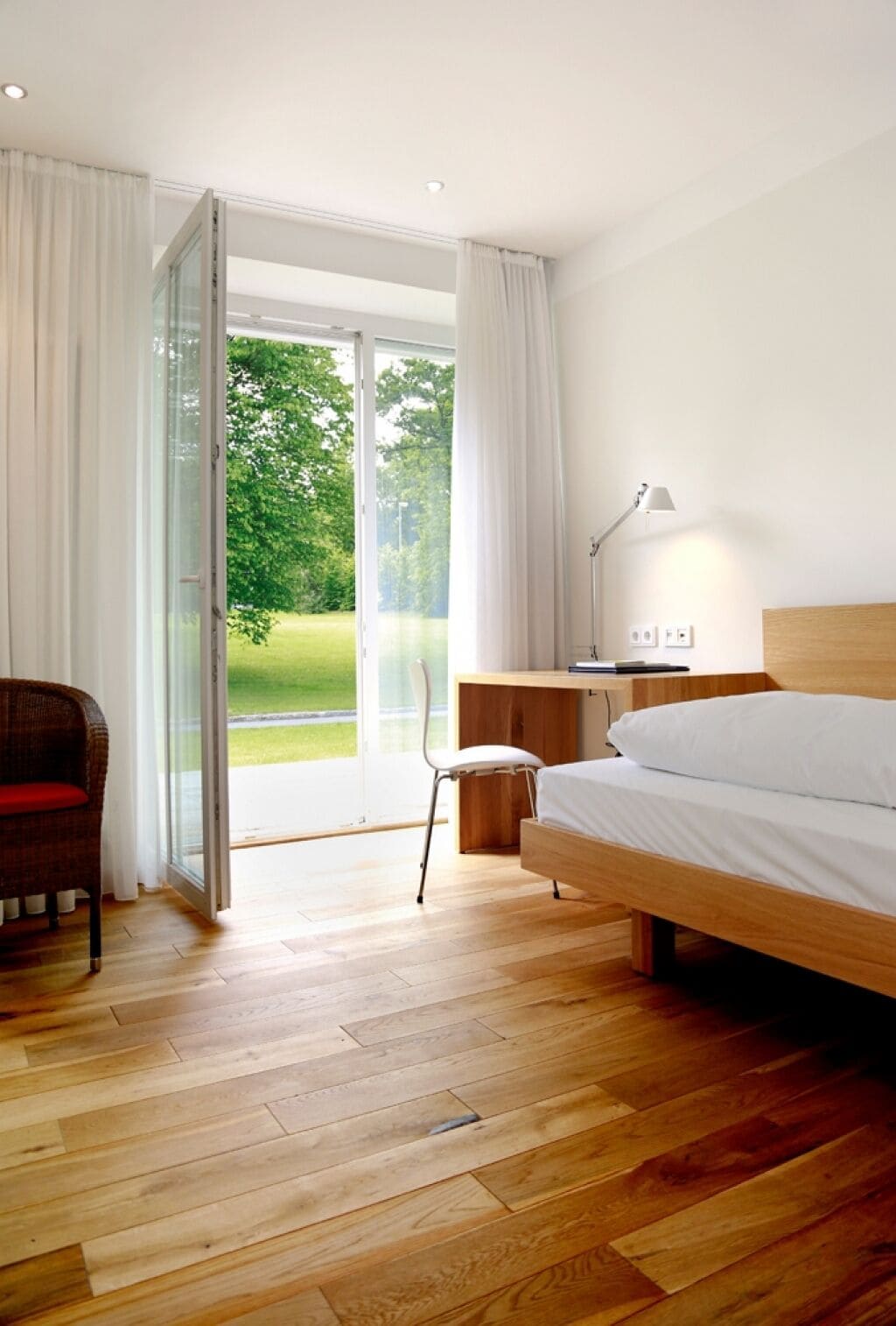 Une chambre d’hôtel moderne avec un plancher en bois, un lit, un bureau, une chaise et une vue sur un paysage verdoyant, visible à travers une porte vitrée.