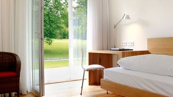 Une chambre d’hôtel moderne avec un plancher en bois, un lit, un bureau, une chaise et une vue sur un paysage verdoyant, visible à travers une porte vitrée.