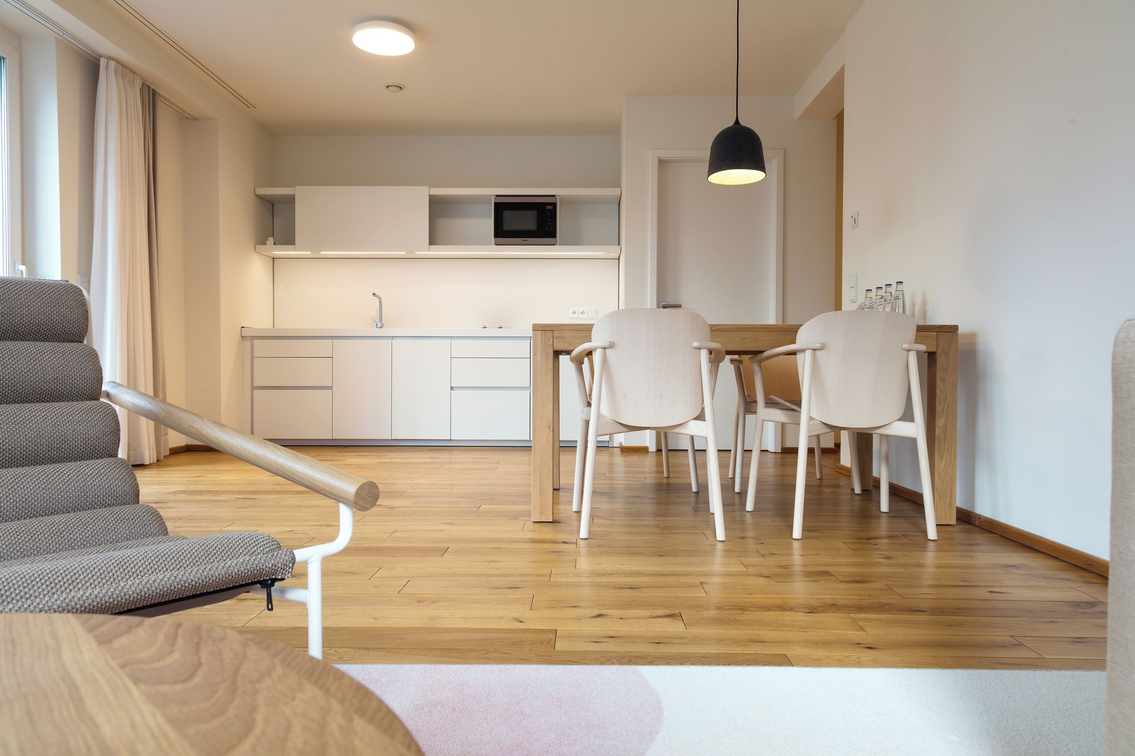A modern living room with a sofa, a dining table with chairs, a kitchen area with a microwave, and a hanging light fixture.