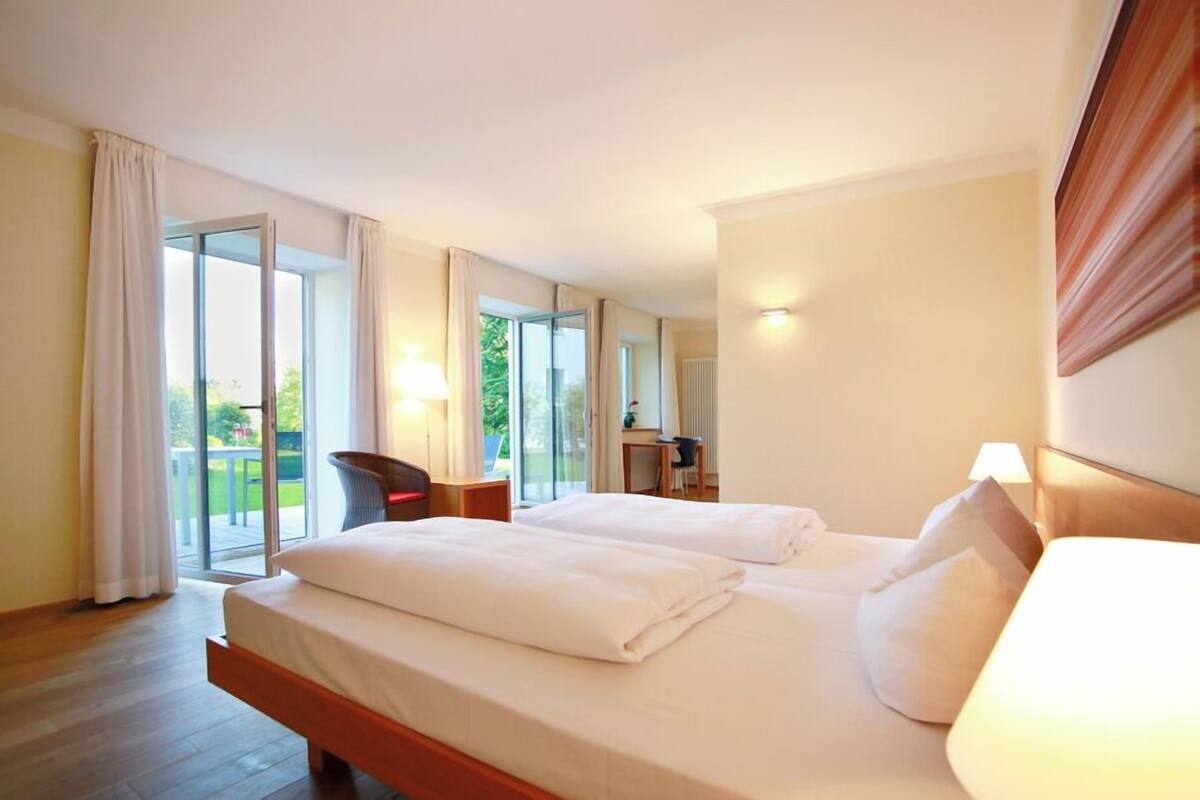 A hotel room with two beds, a chair, a desk, and a view of greenery through large windows.