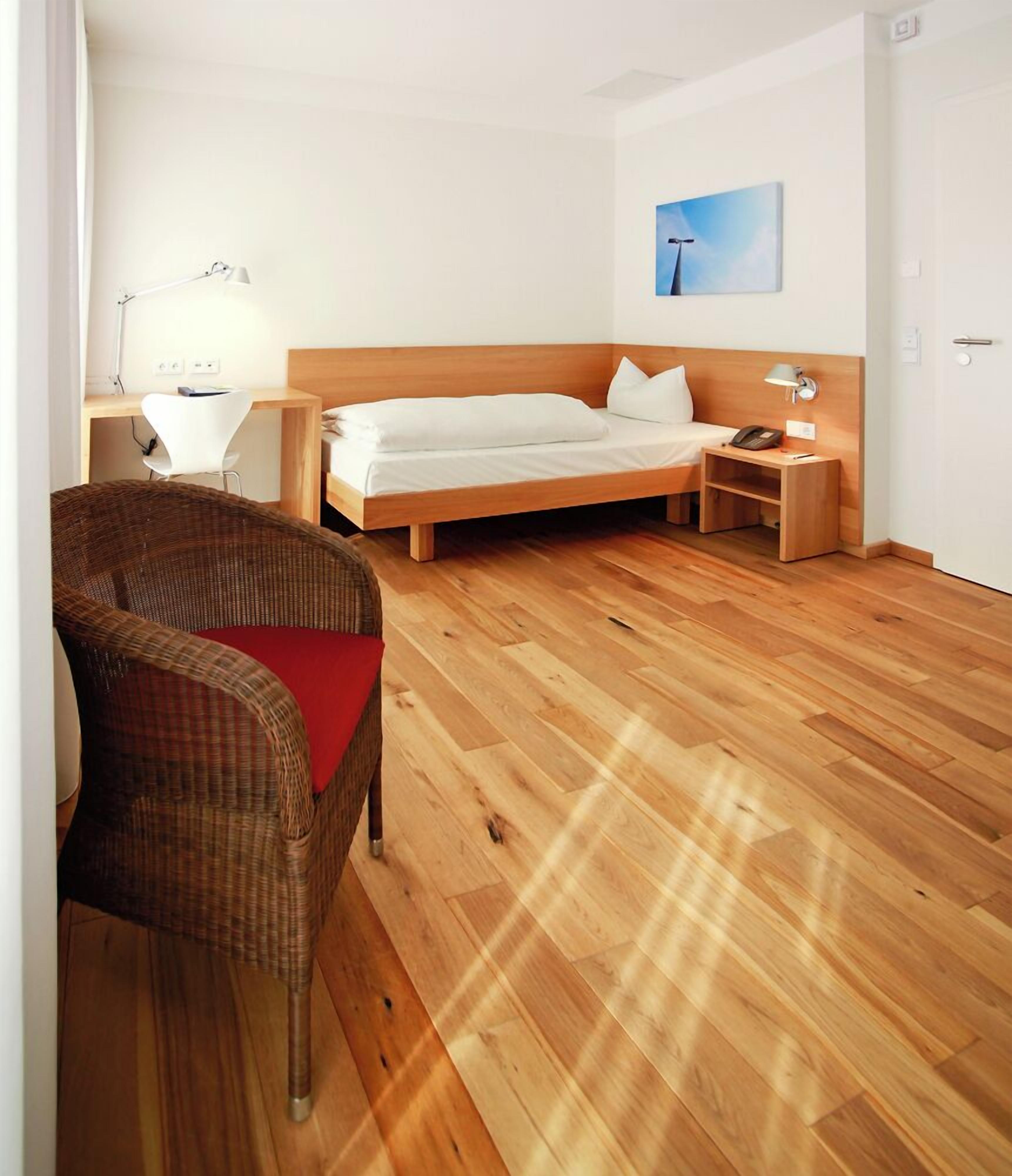 A modern hotel room with a wooden floor, a single bed, a desk with a chair, a small bedside table, and a wall-mounted lamp.
