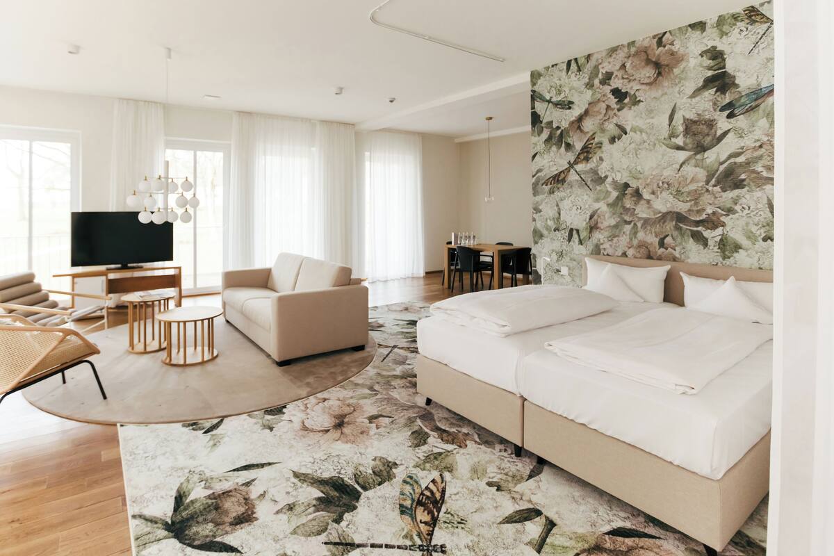 A modern hotel room with a large bed, a sofa, a TV, a dining table, and a floral wall mural.
