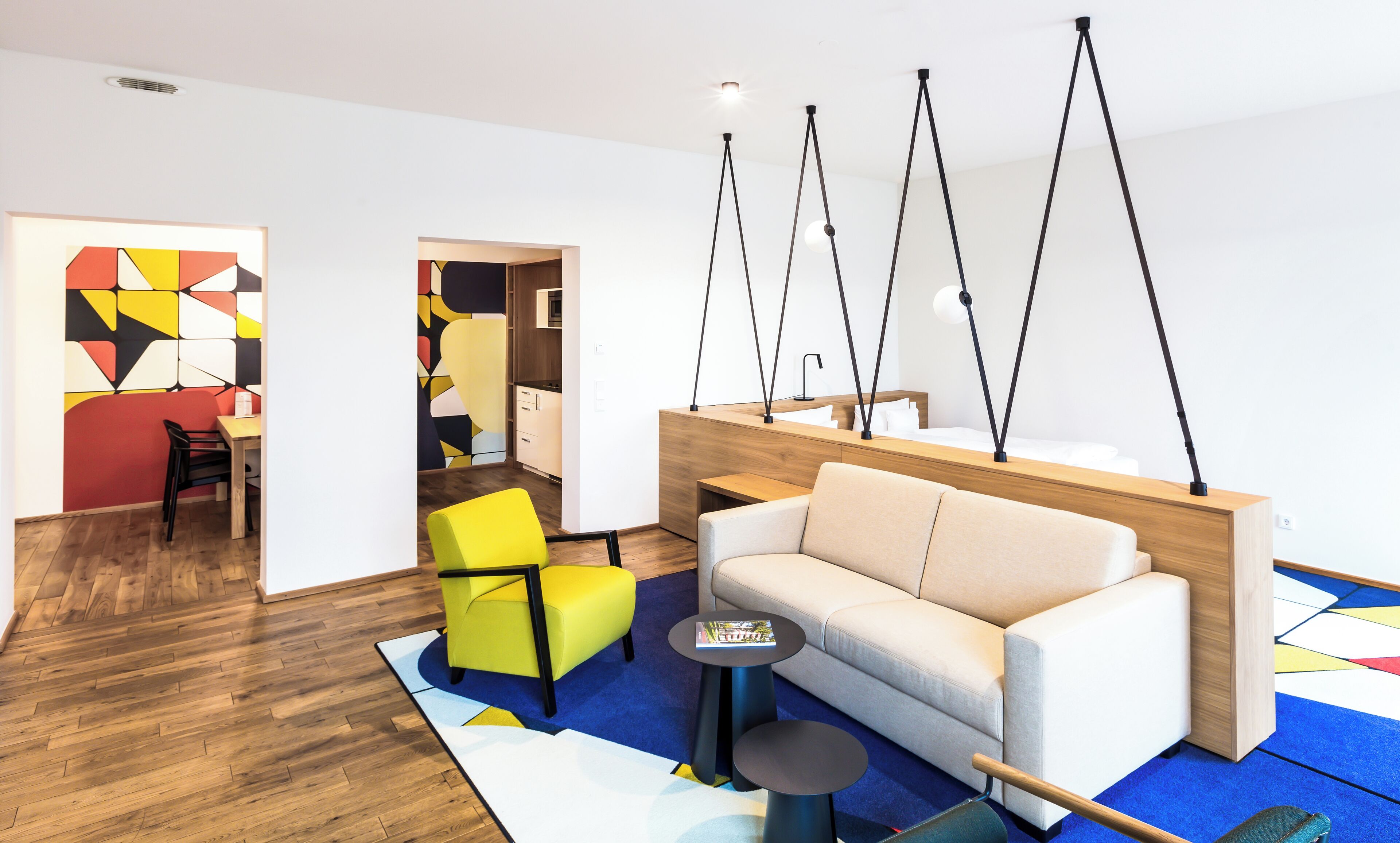 A modern living room with a sofa, a yellow chair, and a coffee table. There is a wooden desk with a lamp and a bed with a white mattress.
