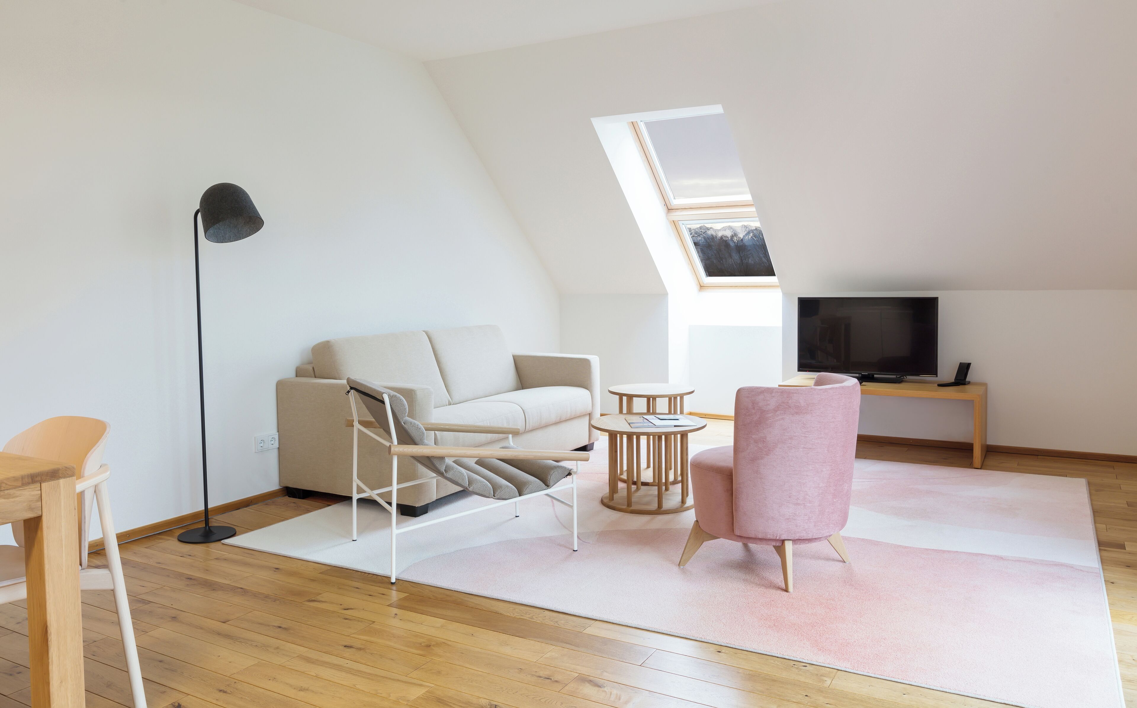 Ein modernes Schlafzimmer im Loft-Stil mit schrägen Wänden, einem großen Dachfenster, einer Couch, einem rosa Sessel, einem kleinen runden Tisch, einer Stehlampe und einem Fernseher.