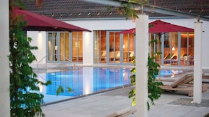 Una piscina con sillones de descanso y sombrillas, rodeada de puertas de vidrio y un suelo de baldosas.