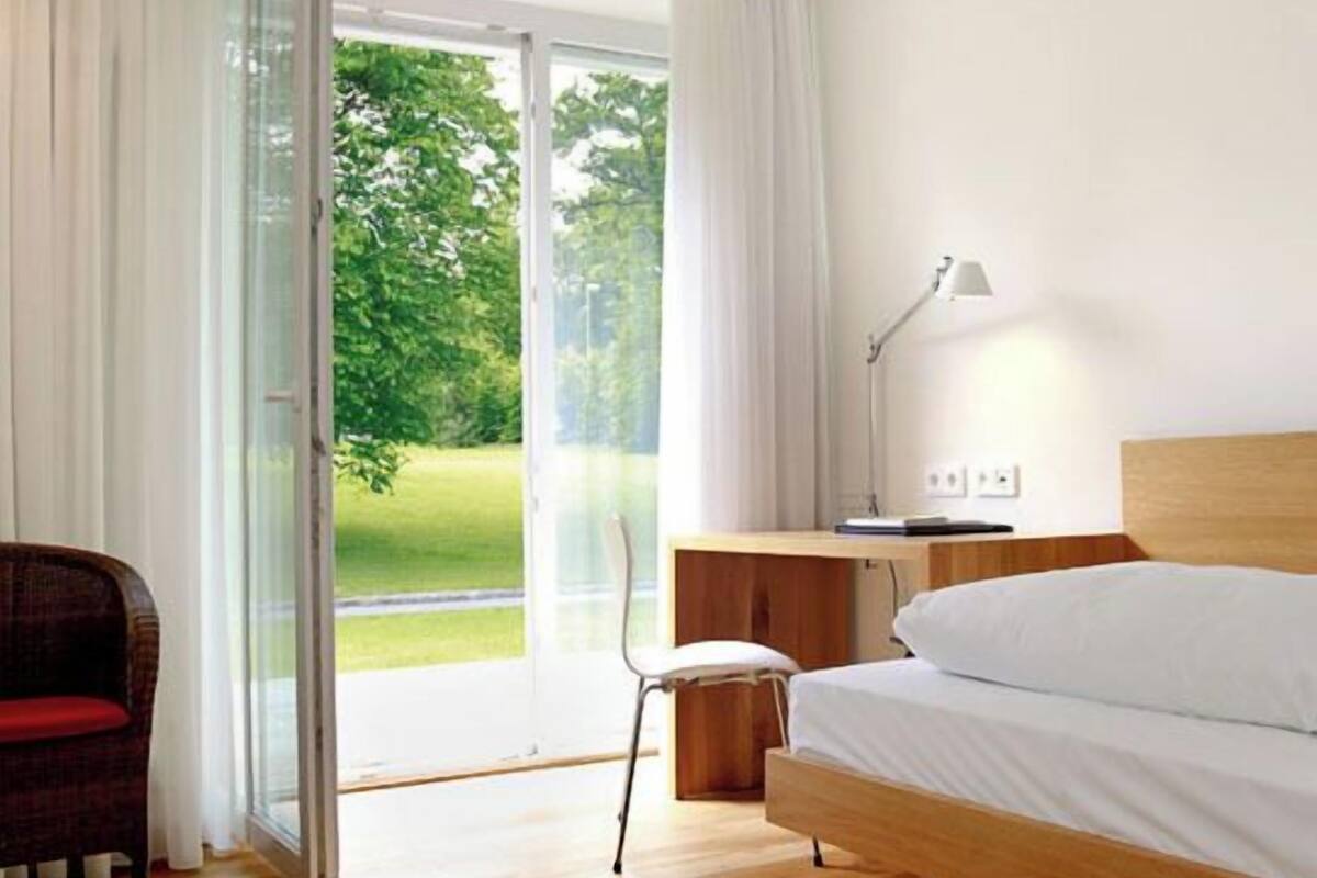 A bedroom with a wooden bed, a desk, a chair, and a view of greenery through a glass door.