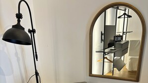 A grey upholstered armchair with wooden armrests, positioned next to a black floor lamp with a curved arm. A large mirror with a gold frame is mounted on the wall above the chair. - Olivuzza 27 (Palermo)
