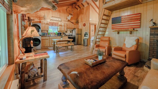 A rustic cabin interior with wooden walls, a dining area, a kitchen, and a living room with a large American flag. - 2Q Cabin near Leesville Lake ~Pets welcome~ (Carrollton)
