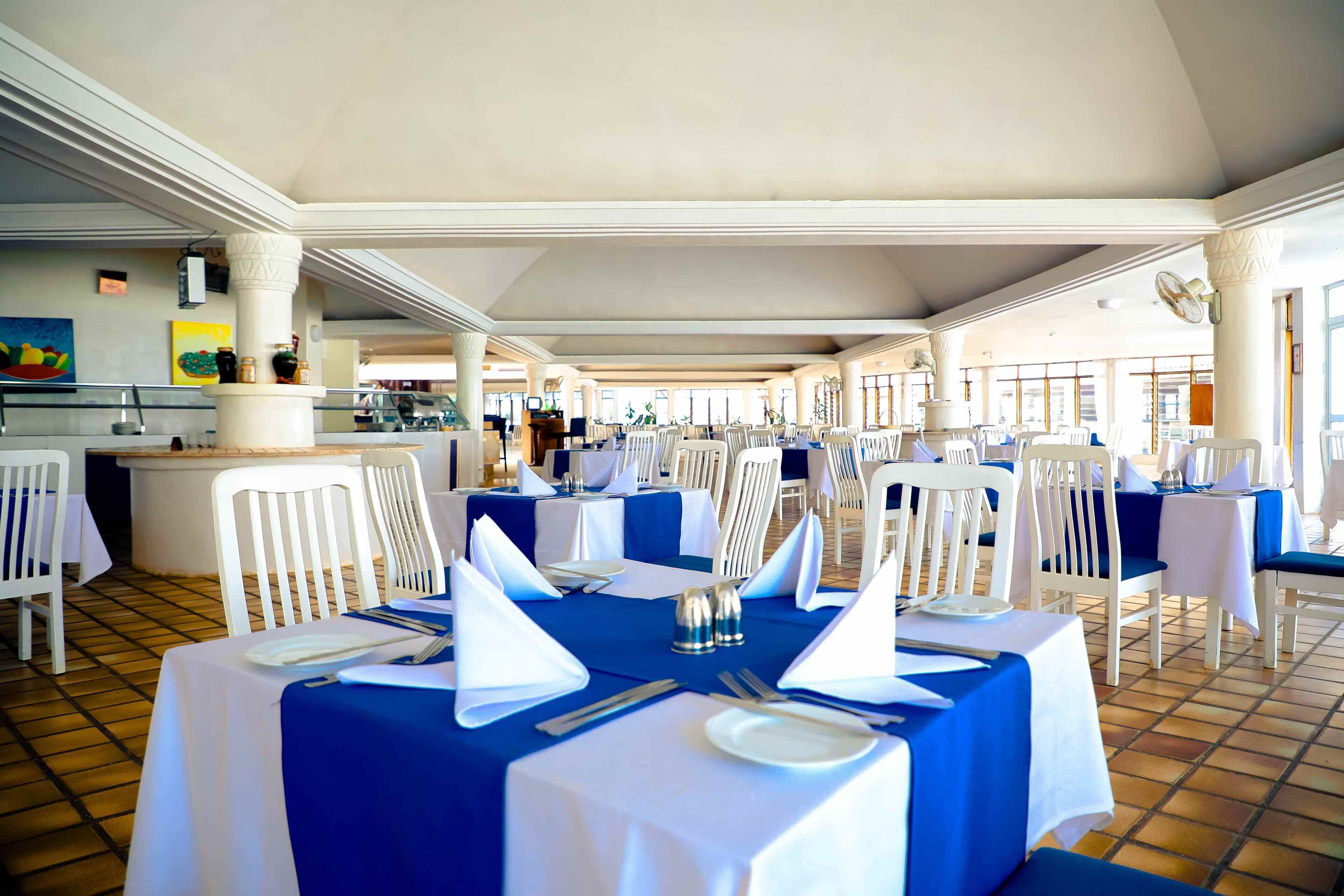 Un comedor con mesas dispuestas para el servicio, sillas blancas y piso de baldosas.