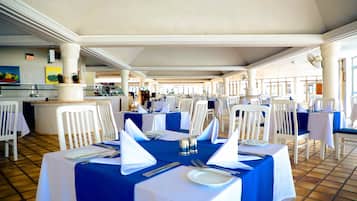 Un comedor con mesas dispuestas para el servicio, sillas blancas y piso de baldosas.