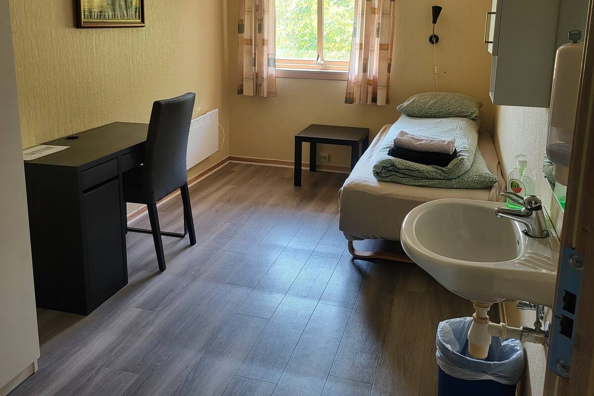 A compact room with a bed, a desk, a chair, a window with curtains, a sink, and a trash bin.