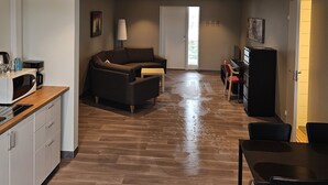 A spacious living area with a sofa, coffee table, and a dining table with chairs. There is a kitchen area with a microwave and coffee maker. The room has wooden flooring and a large window. - Solvoll (Tønsberg)