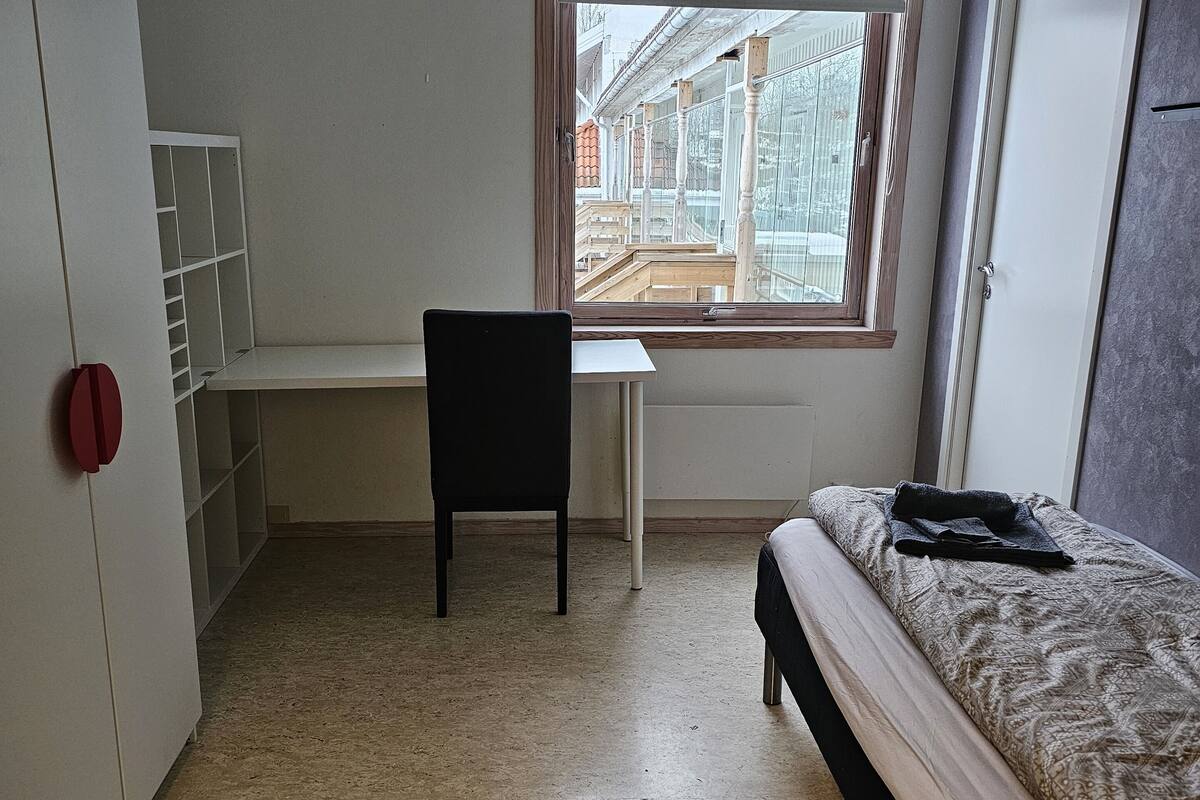 A room with a bed, a desk, and a chair. There is a window with a view of buildings outside.