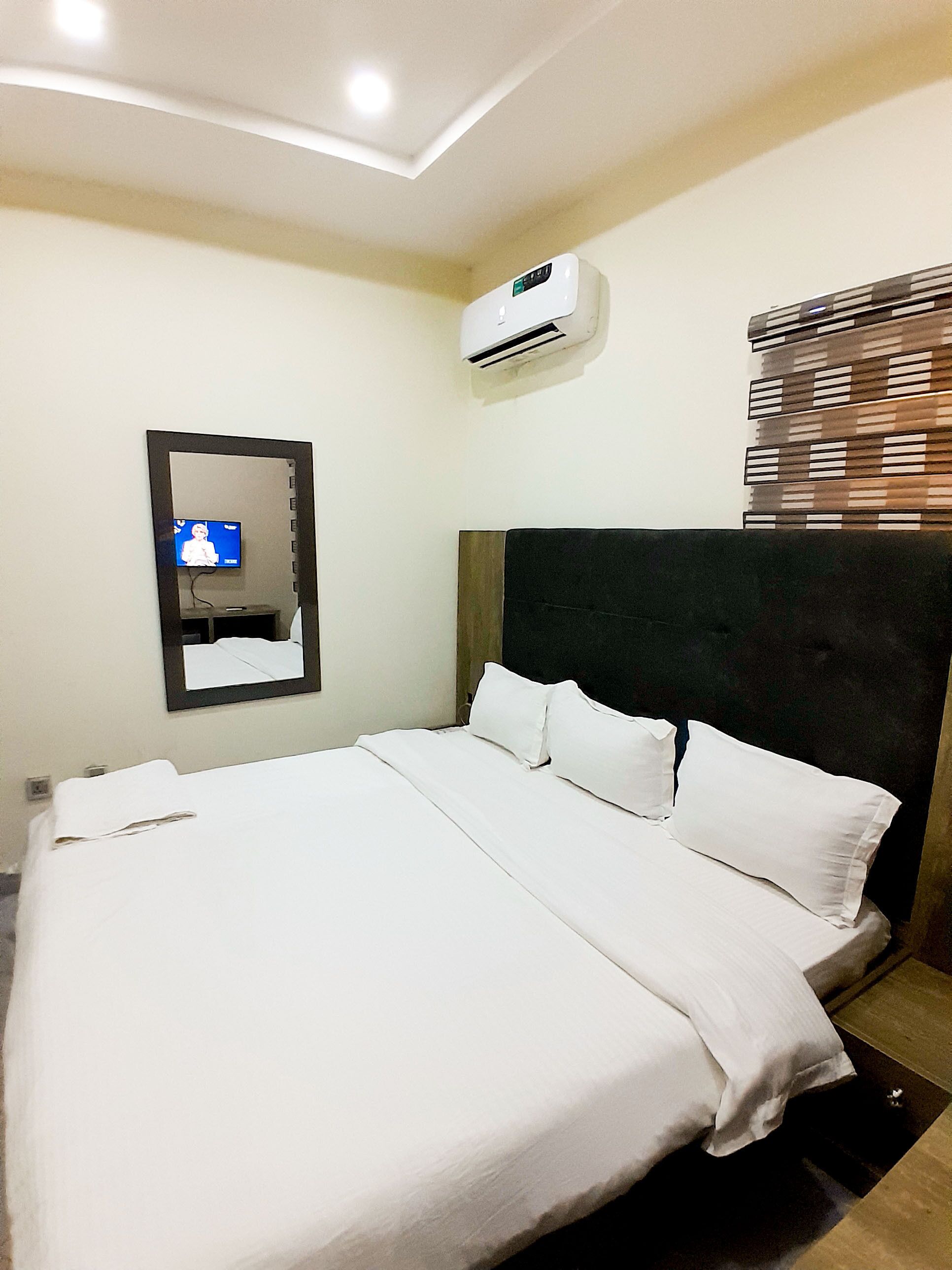 A hotel room with a large bed, a mounted air conditioner, a flat-screen TV, and a window with blinds.