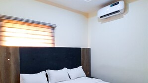 A hotel room with a large bed, a nightstand, a wall-mounted air conditioner, and a window with blinds. - Come Inn Hotel (Abuja)
