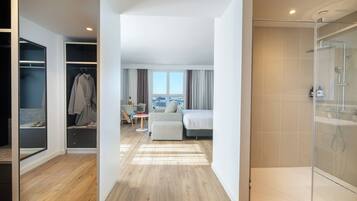 A modern hotel room with a walk-in closet, a bed, a seating area with a view, and a glass-enclosed shower.