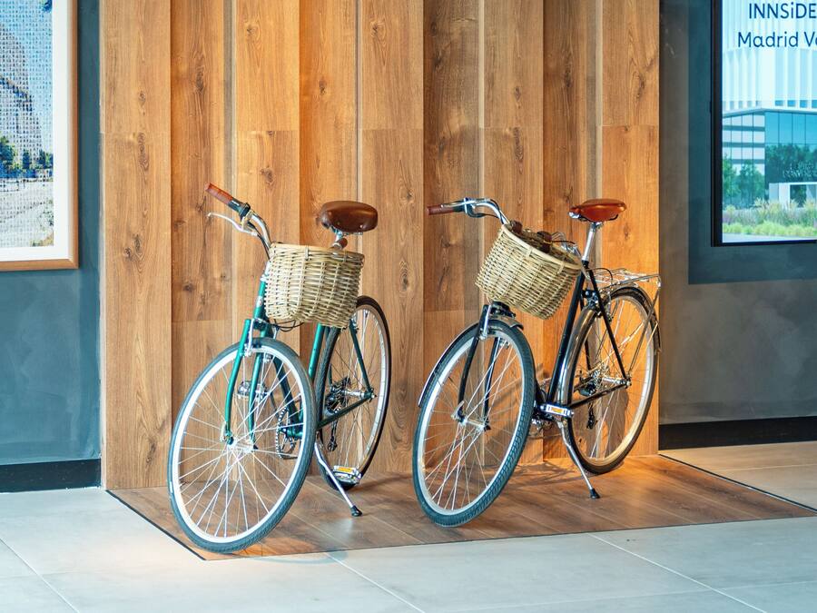 Two bicycles with baskets against a wooden wall, a framed picture, and a large window with a view of a building.