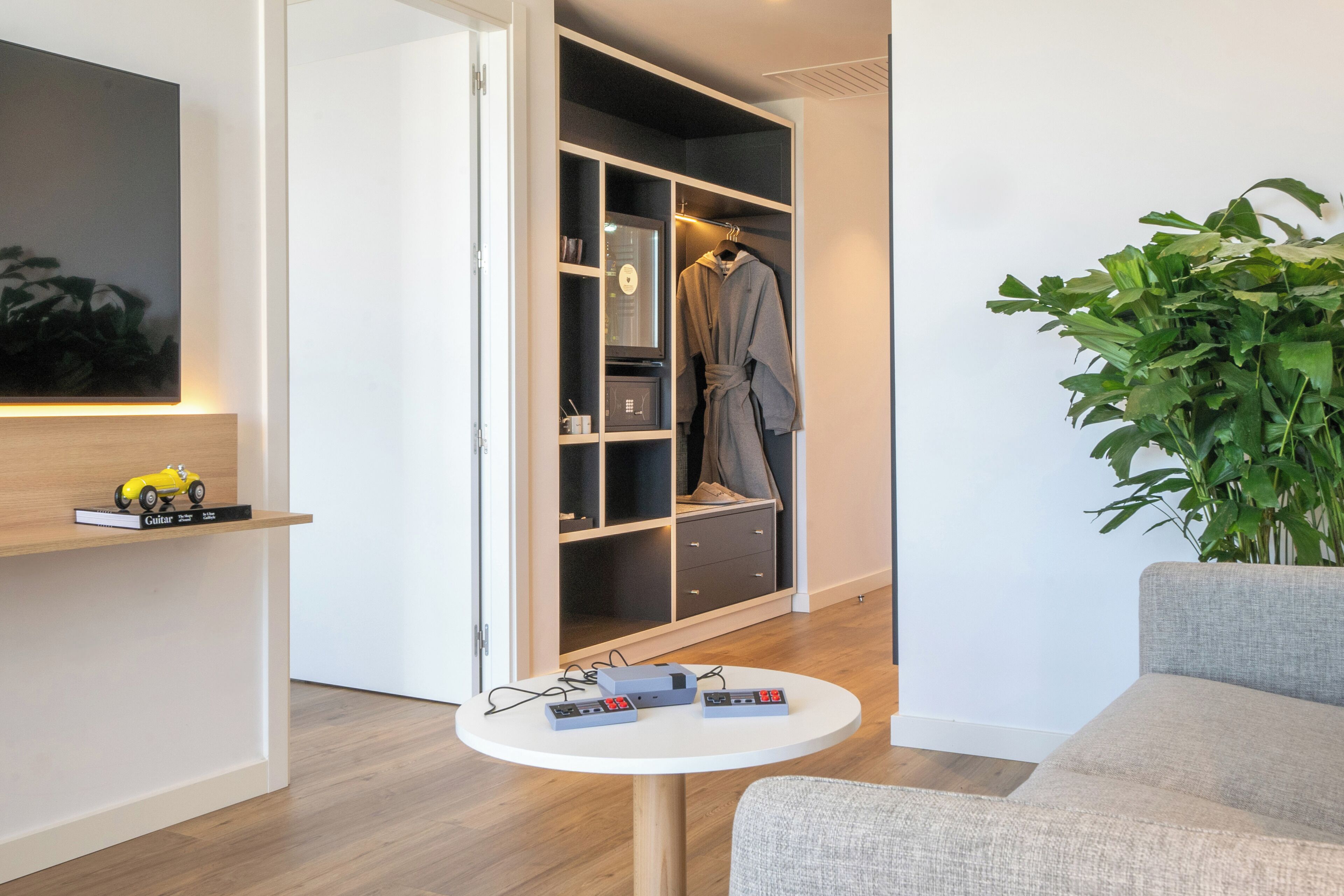 Una habitación moderna con un sofá gris, una mesa redonda con dispositivos electrónicos, un armario con bata y una planta en maceta.