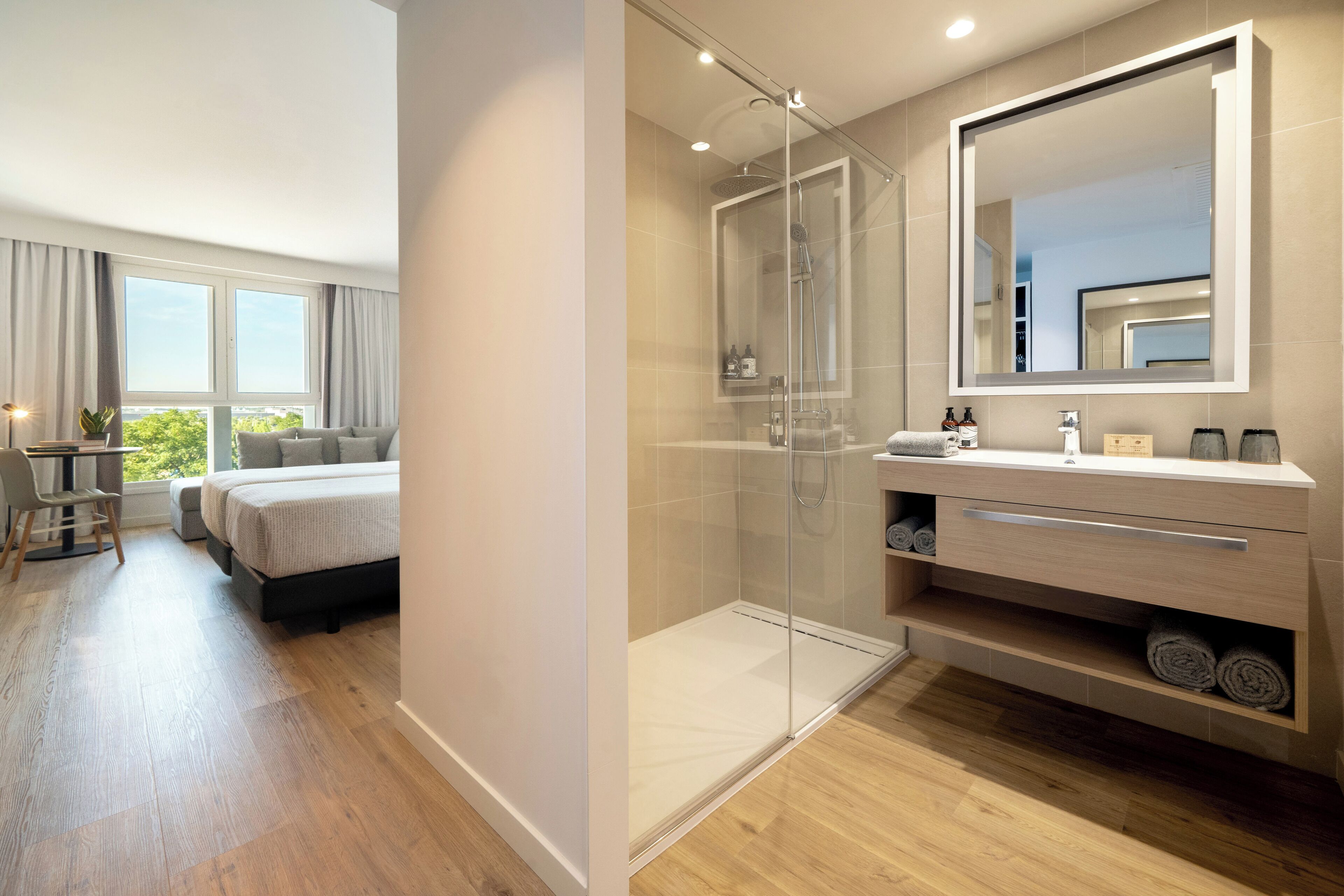 A modern bedroom with a glass-enclosed shower, a bedside table, a bed, and a large mirror above the vanity.