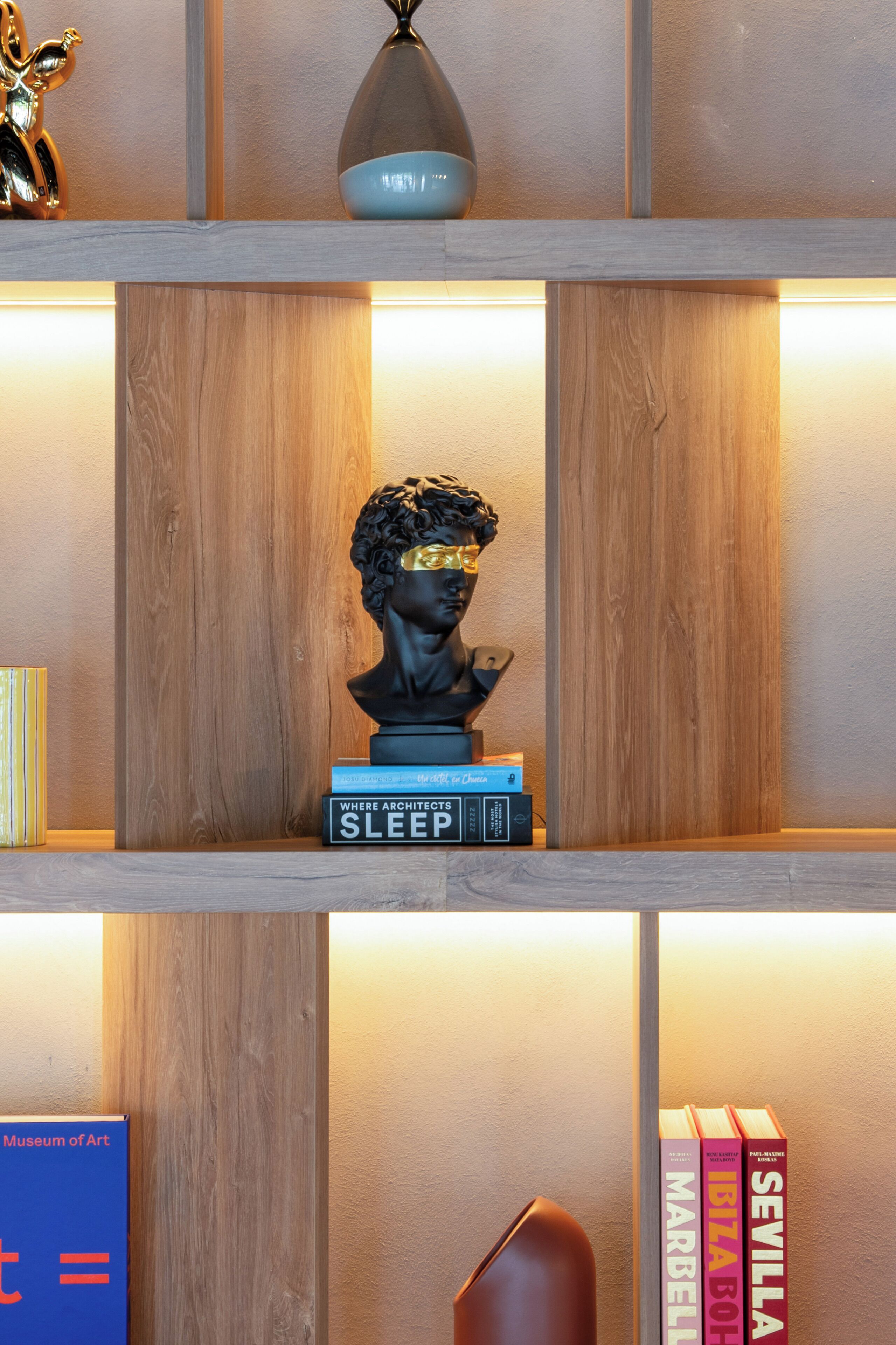 Una estantería con libros, un busto y una lámpara.