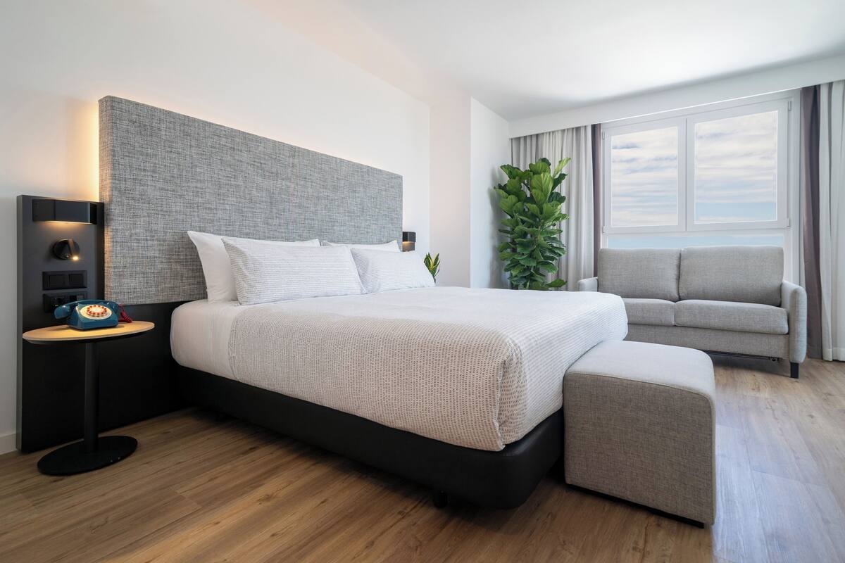 A modern hotel room with a large bed, a side table with a telephone, a grey upholstered chair, and a large window with a view.