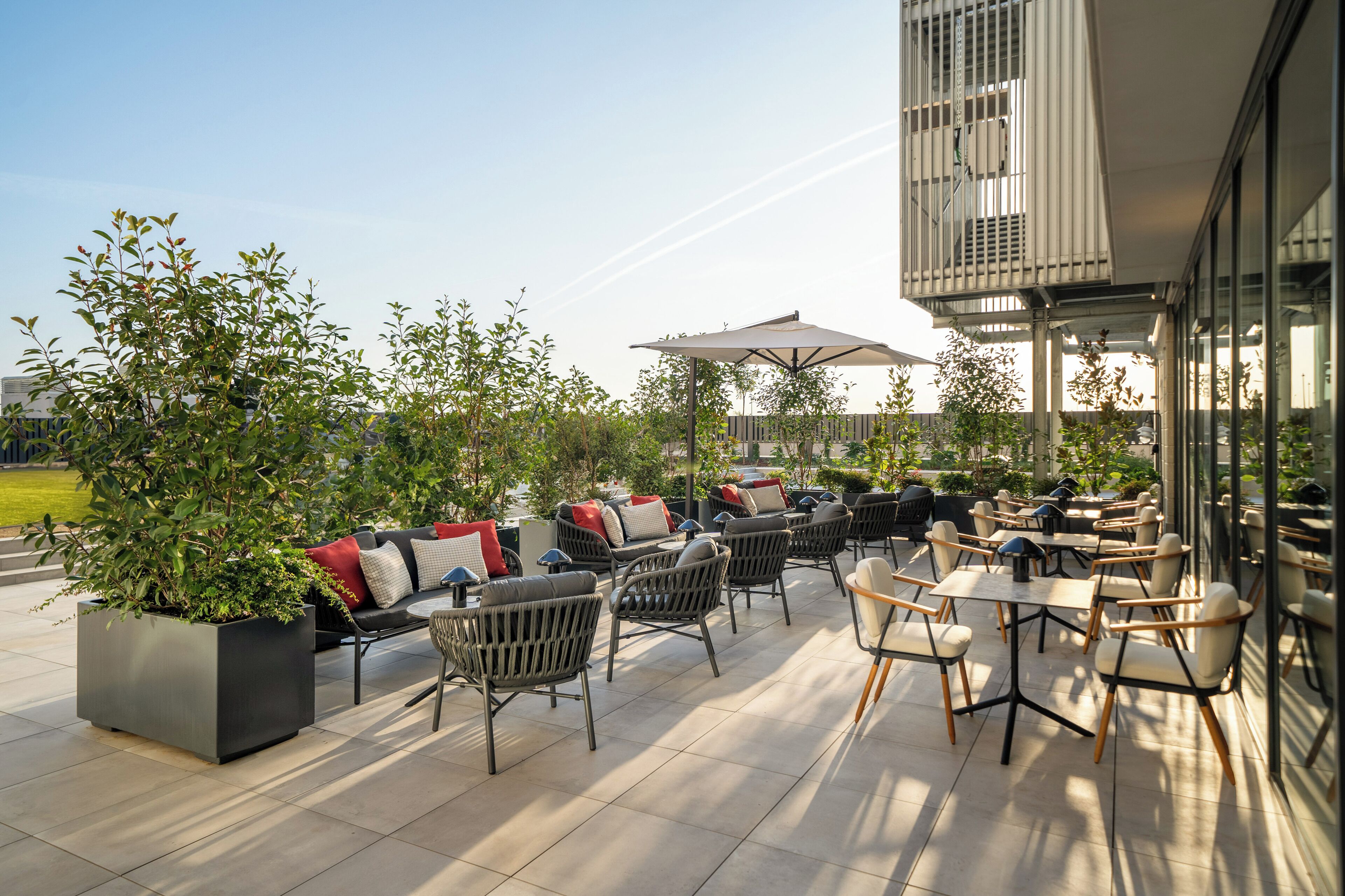 Un patio al aire libre con asientos y mesas, rodeado de follaje y vistas de un edificio.