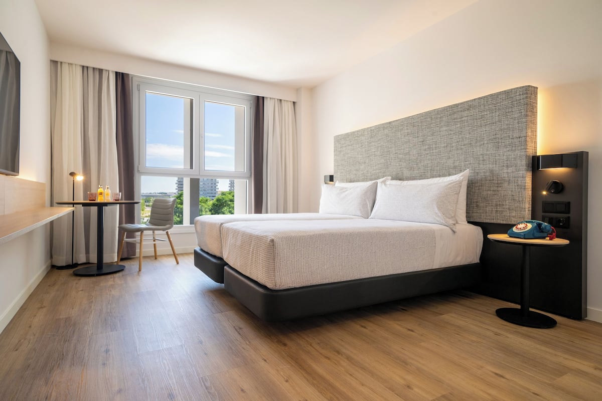 A modern hotel room with a large bed, a desk with a chair, and a view of the city through large windows.