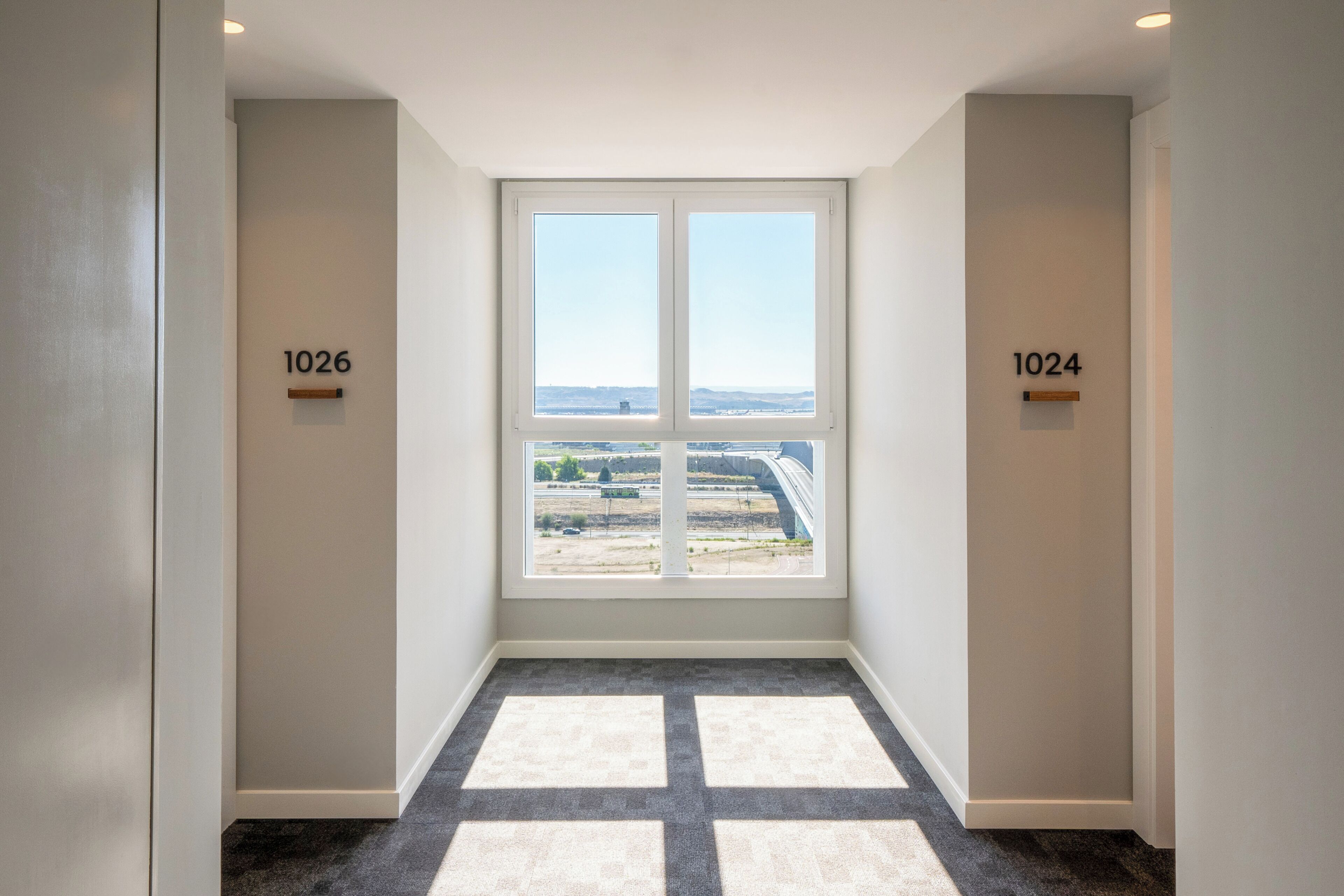A hallway with a large window, carpeted floor, and apartment numbers 1026 and 1024 on the walls.