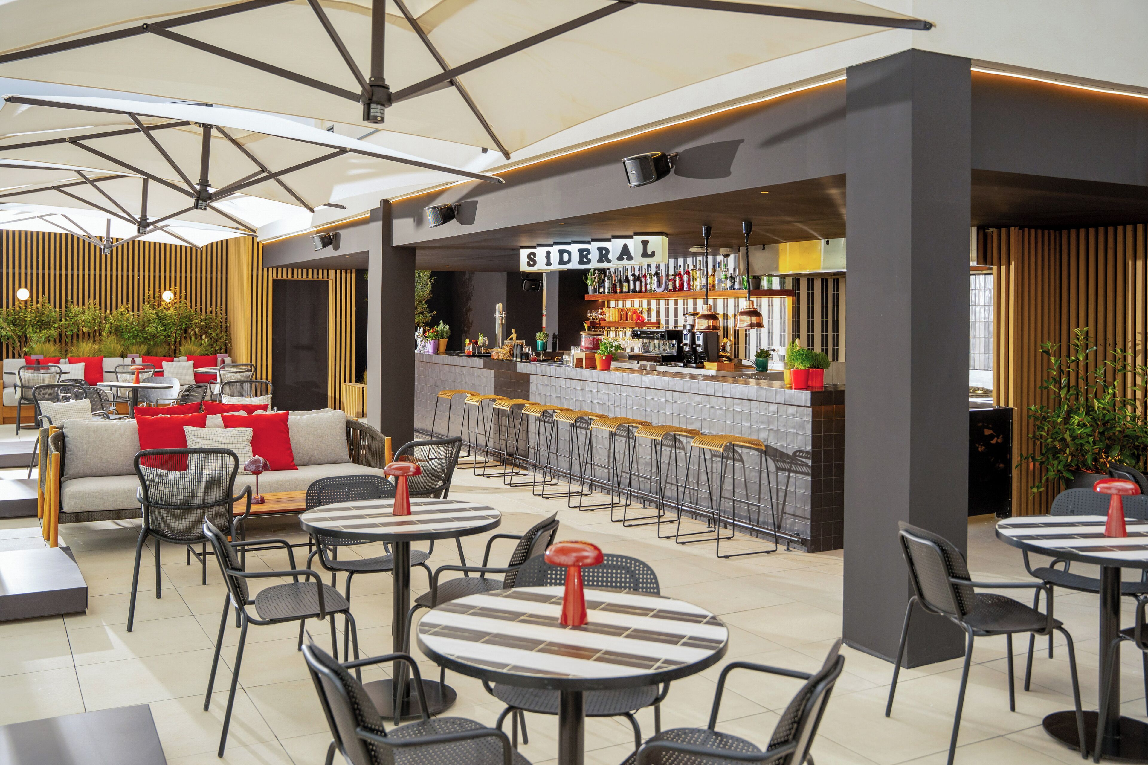 A modern outdoor seating area with tables and chairs, a bar with a sign reading "SIDERAL," and potted plants.