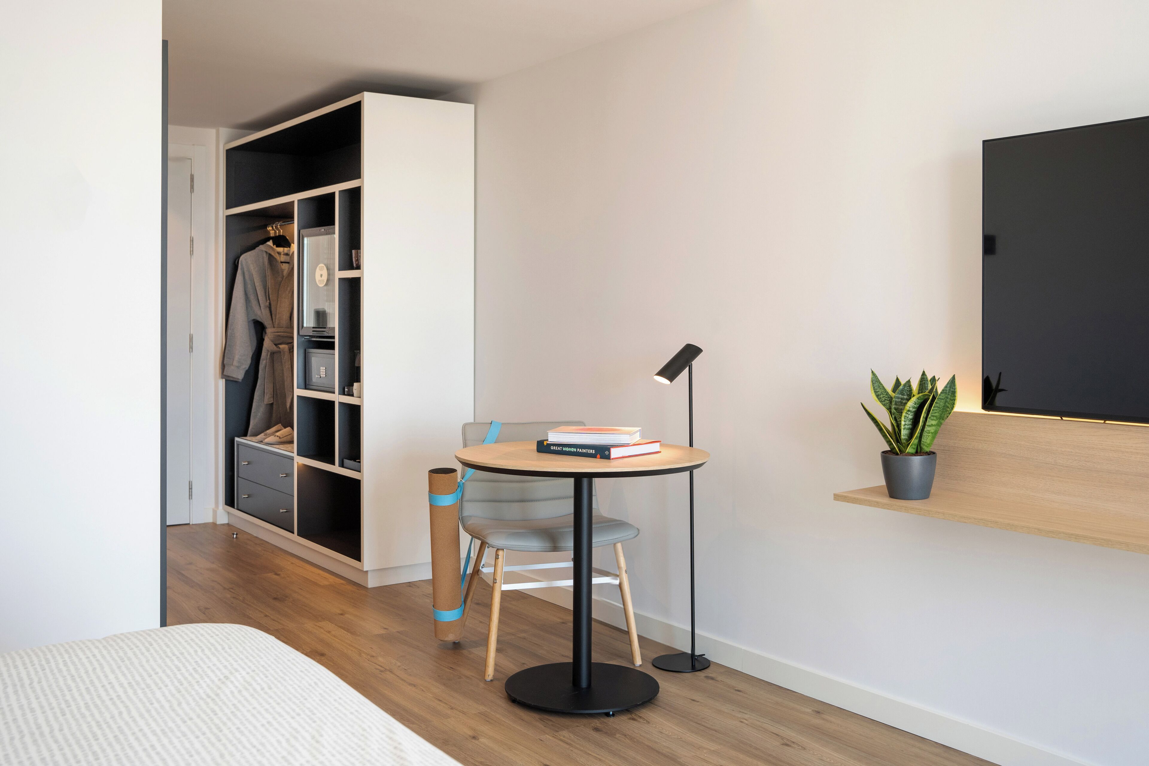 Un dormitorio con cama, armario, una pequeña mesa redonda con libros, lámpara de pie y planta en maceta.