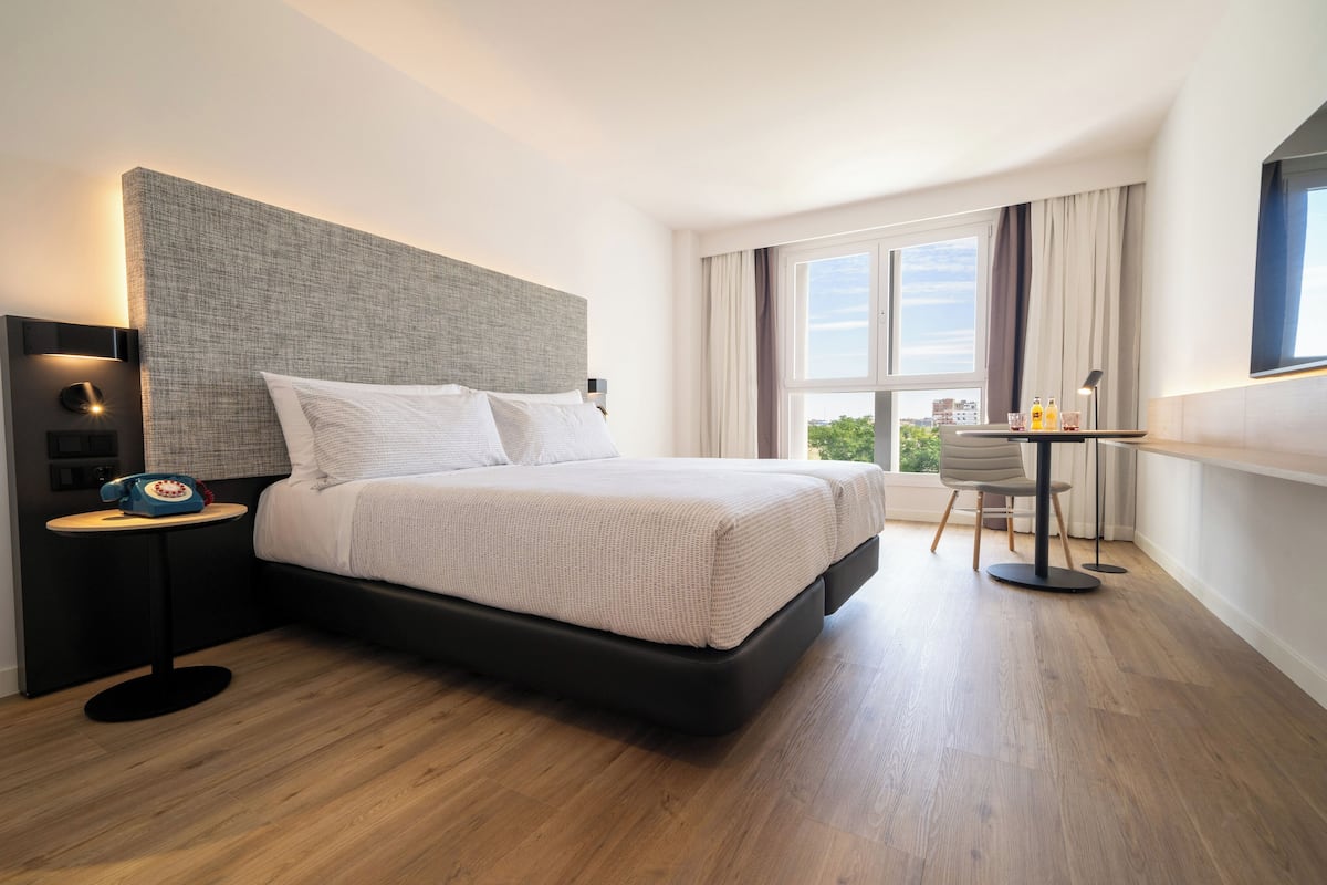 A modern hotel room with a large bed, a small desk, and a view of the city through the window.