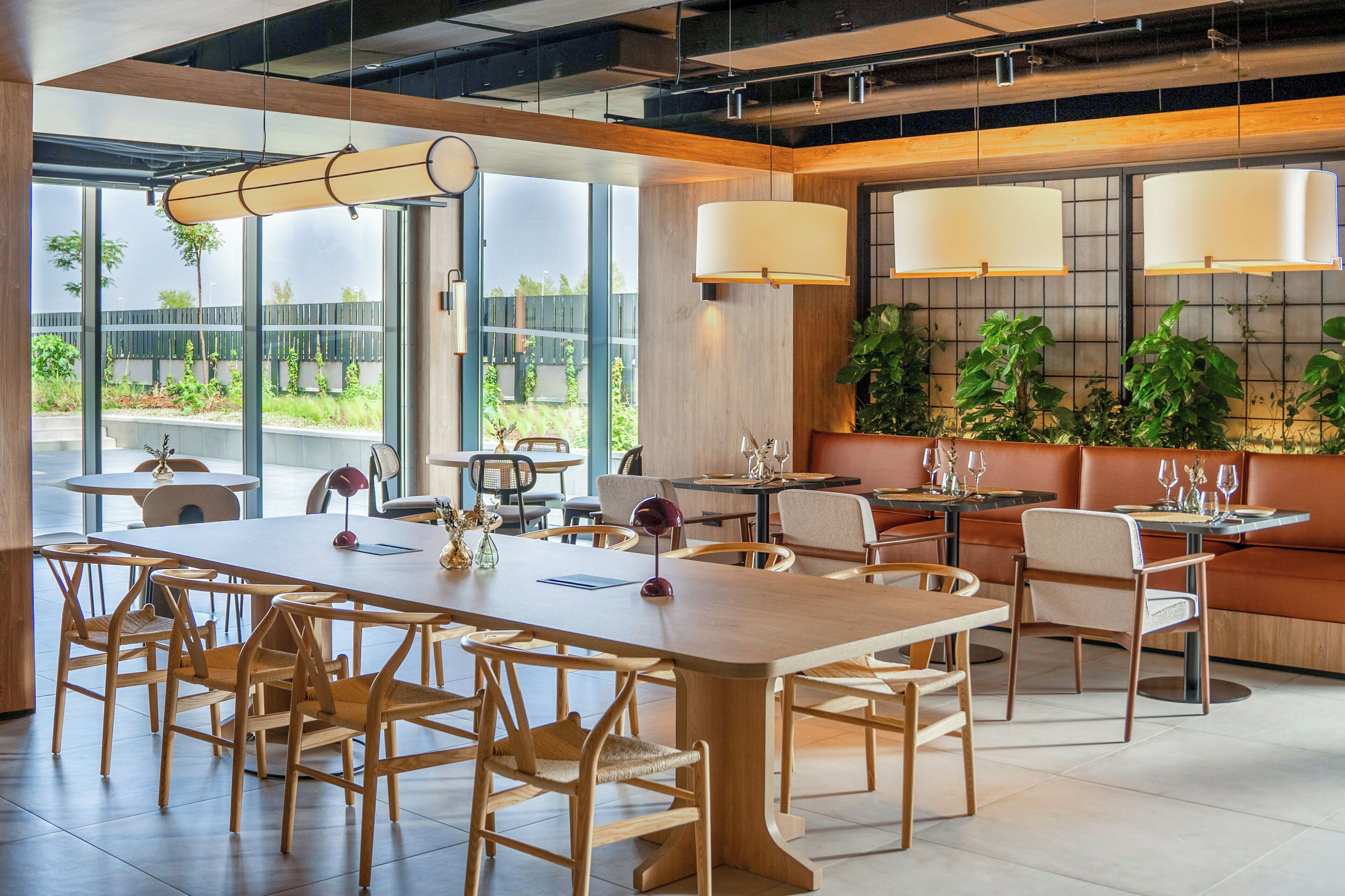 A modern dining area with wooden tables and chairs, large windows, and indoor plants.