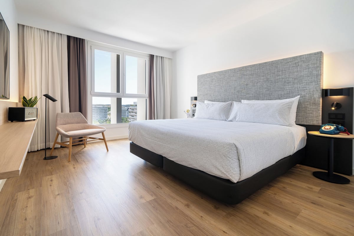 A modern hotel room with a large bed, a desk, a chair, and a view of the city through the window.