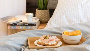 A neatly made bed with a light blue blanket, a bedside table with a book titled 'EVOLUTION', a small potted plant, and a tray with a sandwich and a bowl of fruit.