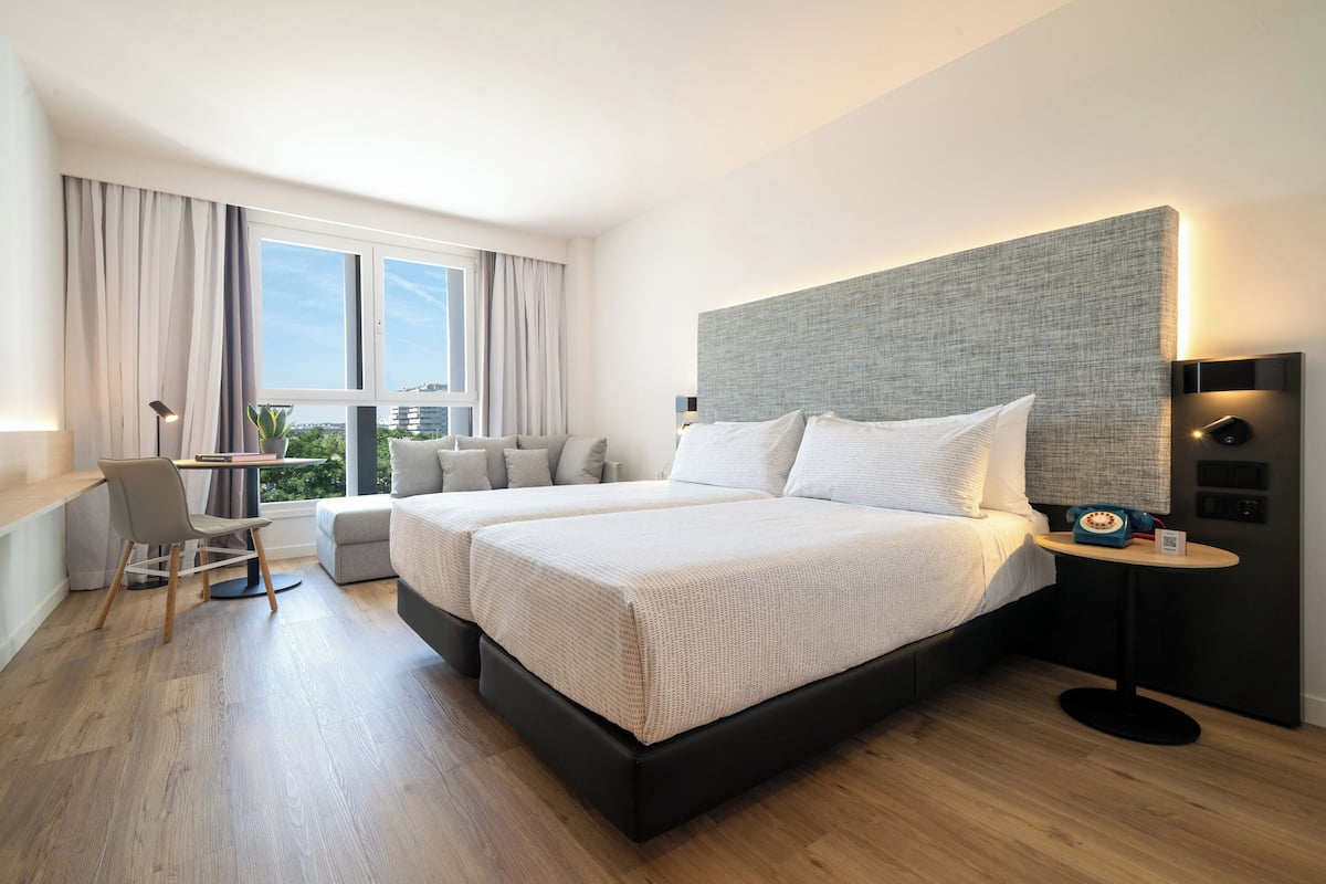 A modern hotel room with two beds, a desk, and a view of the city.