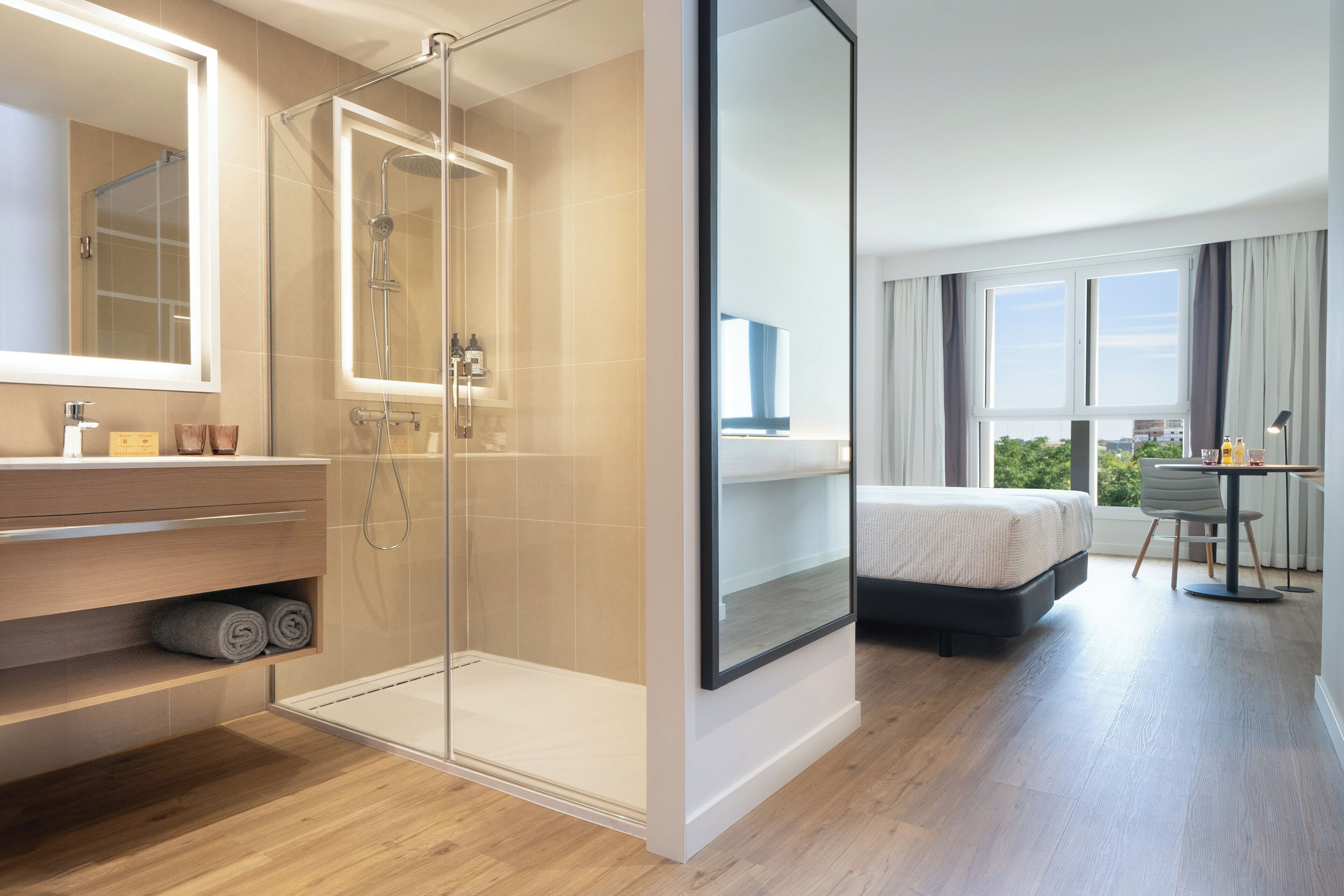 A modern hotel room with a glass-enclosed shower, a large bed, a desk with a chair, and a view of the outdoors.