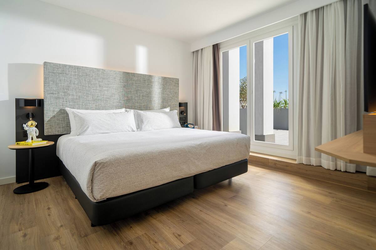 A modern hotel room with a large bed, a nightstand, a lamp, and a view of the outdoors through a large window.