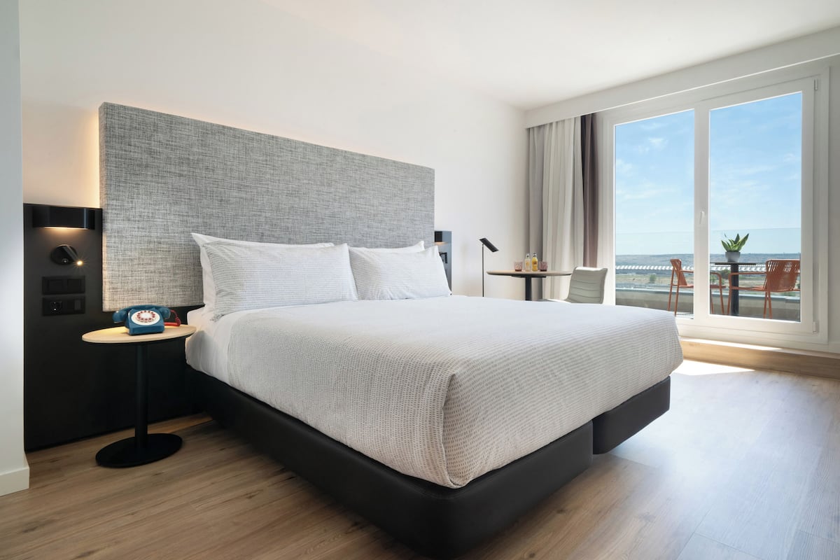 A modern hotel room with a large bed, a bedside table with a lamp, a small table with a plant, and a view of the sea through large windows.