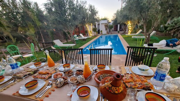 An outdoor dining area with a pool, green chairs, and a table set with plates, cutlery, and various dishes. - Manzal Lahbab (Marrakech)