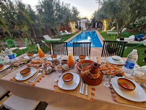 An outdoor dining area with a pool, green chairs, and a table set with plates, cutlery, and various dishes. - Manzal Lahbab (Marrakech)