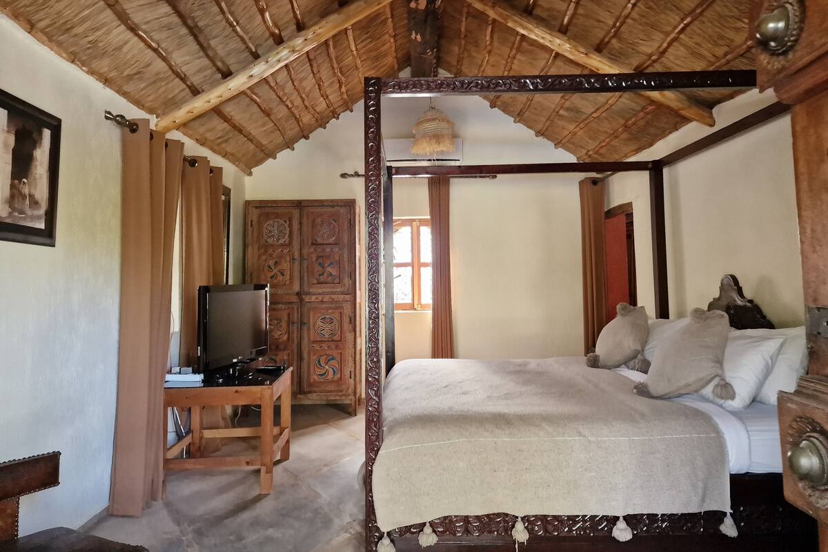 A traditional room with a wooden ceiling, a bed with a canopy, a television on a wooden table, and a wardrobe with intricate designs.