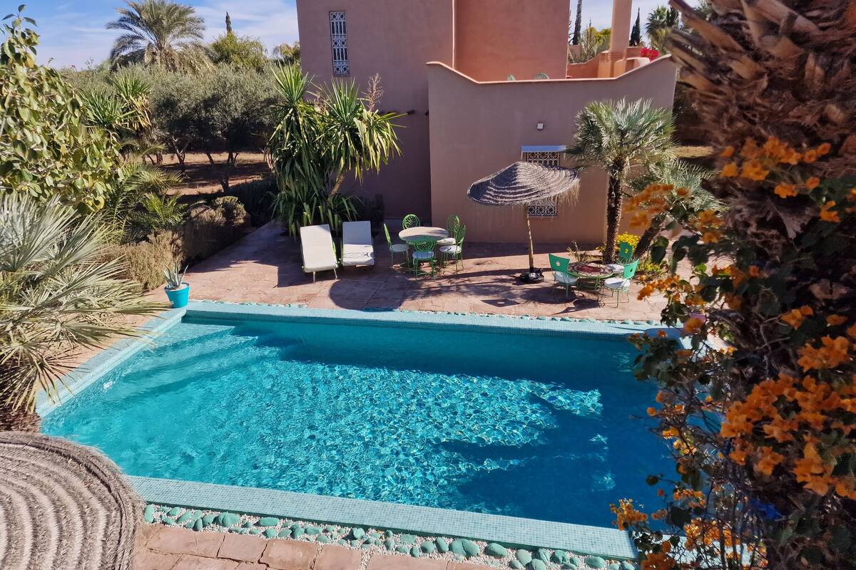 A swimming pool with lounge chairs and a table, surrounded by palm trees and a building with a tiled roof.